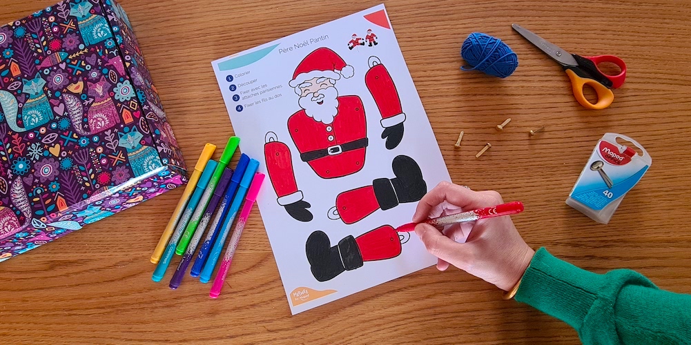 A hand colouring in the sections of a Santa puppet with felt pens lay on the desk